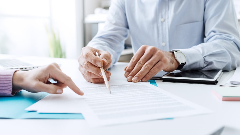 Desk with paperwork, a hand pointing, another hand pointing with a pencil.