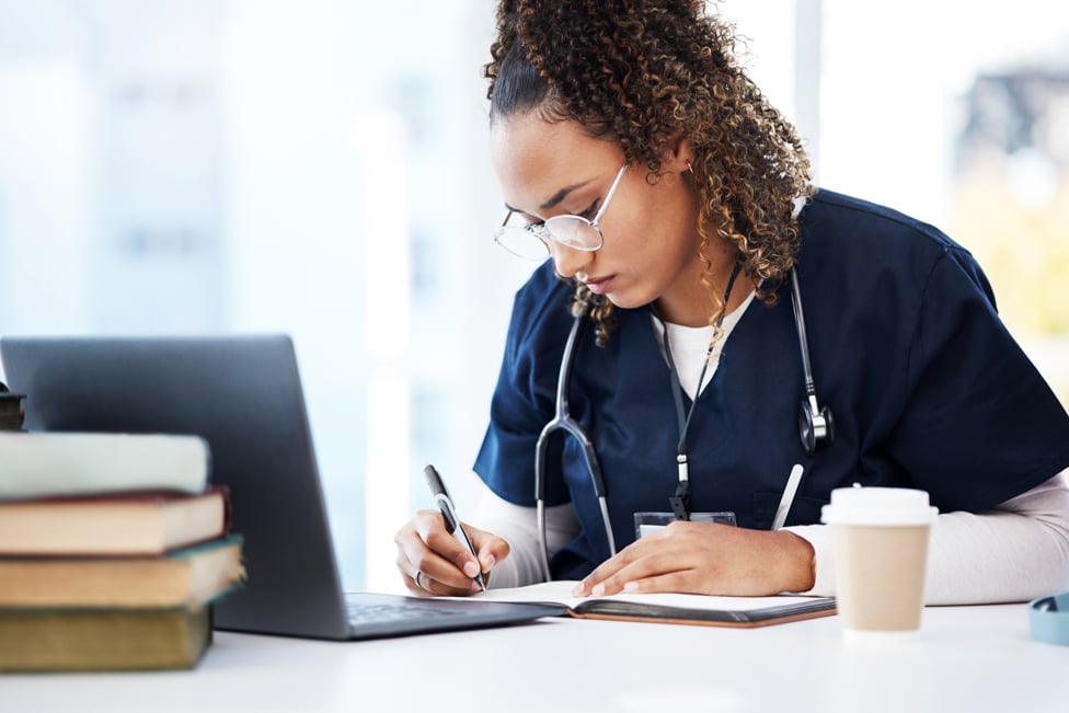 Nurse sitting at table writing notes with a coffee nearby. 