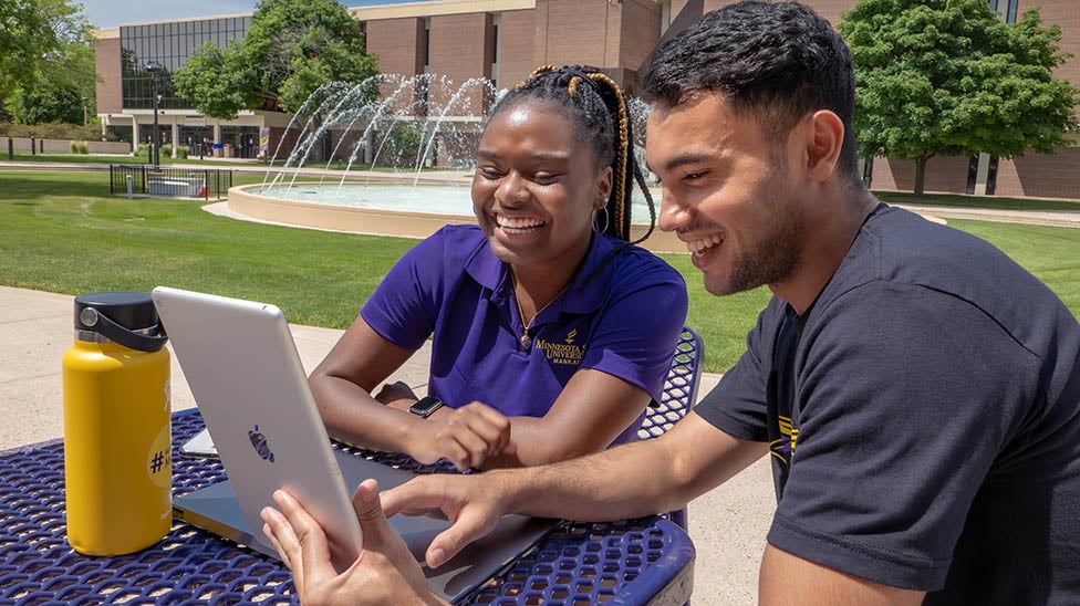 two students sitting outside near the fountain at one of the tables. They are looking at the laptop and smiling.