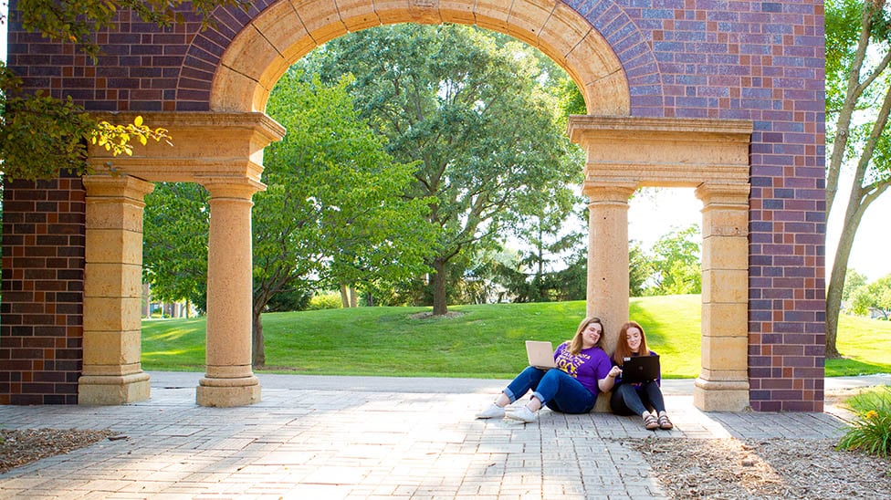 two female students sitting outside on campus under the arch and working on their laptops