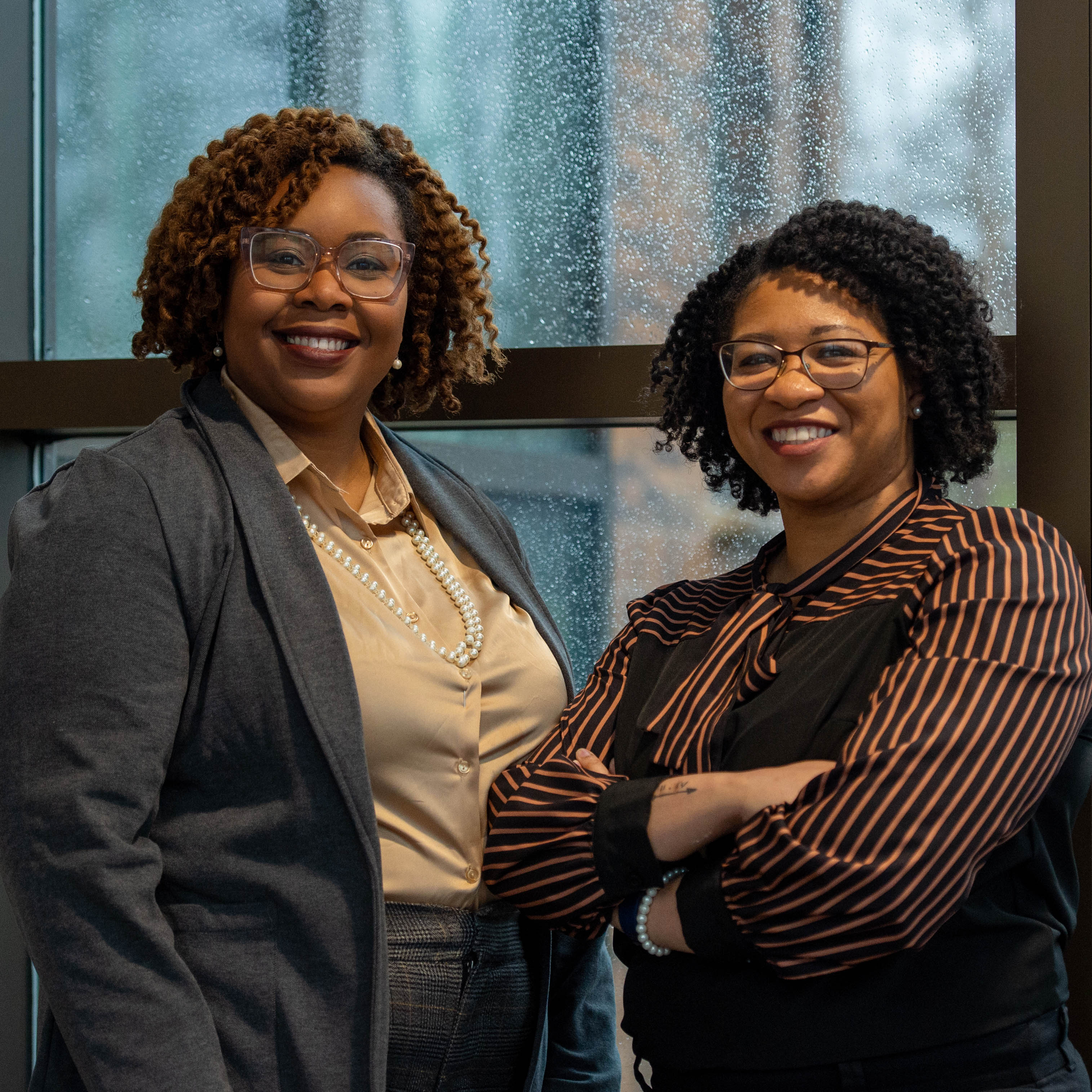 Best friends Felicia Smith and Chalandra Gooden in professional attire smiling together in the hallway of their work building.