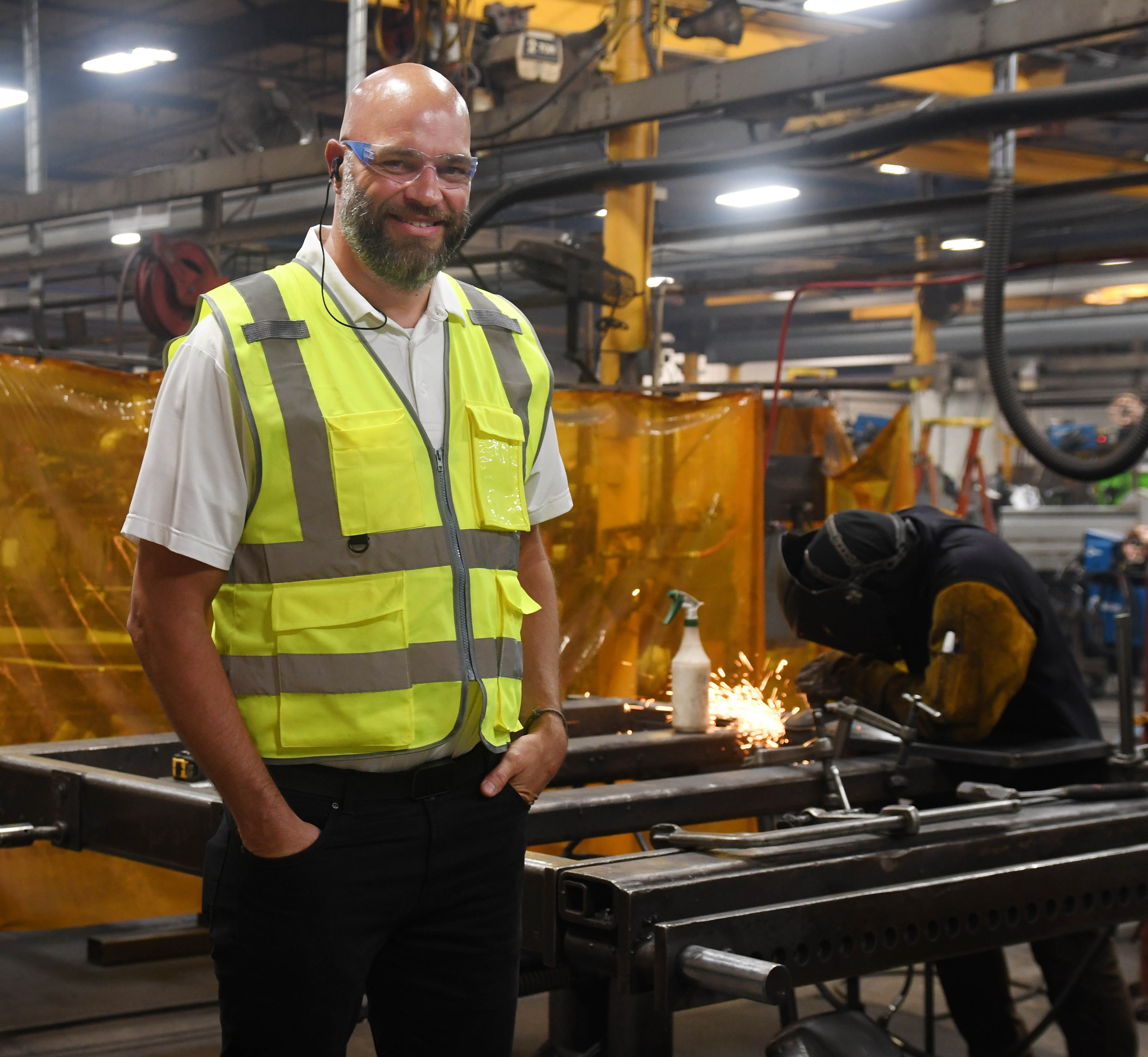 Ben Schmitt wearing a safety vest and goggles standing on a factory floor with welders in the background