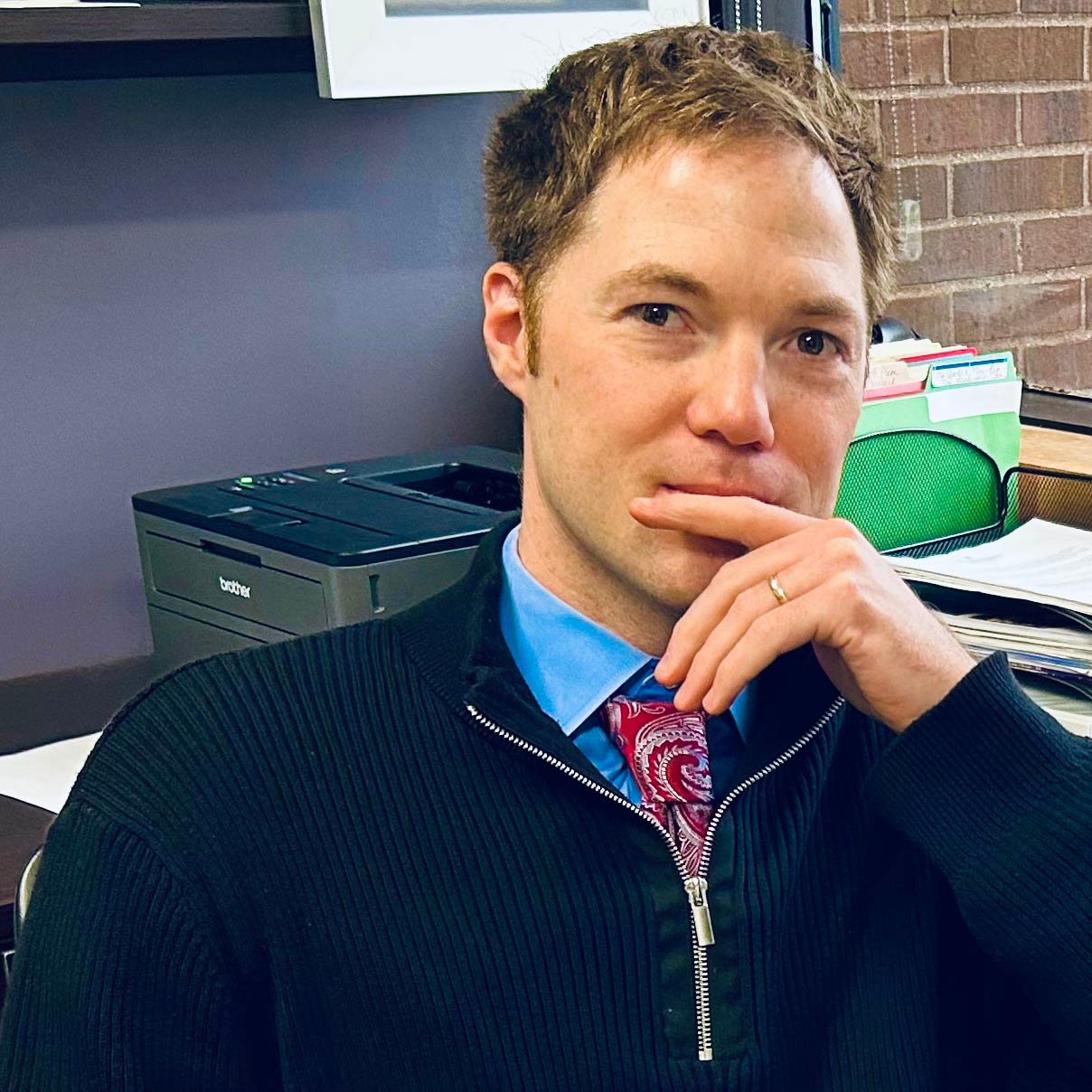 Travis Thul sitting at his desk in his office on campus. He is smiling at the camera. 
