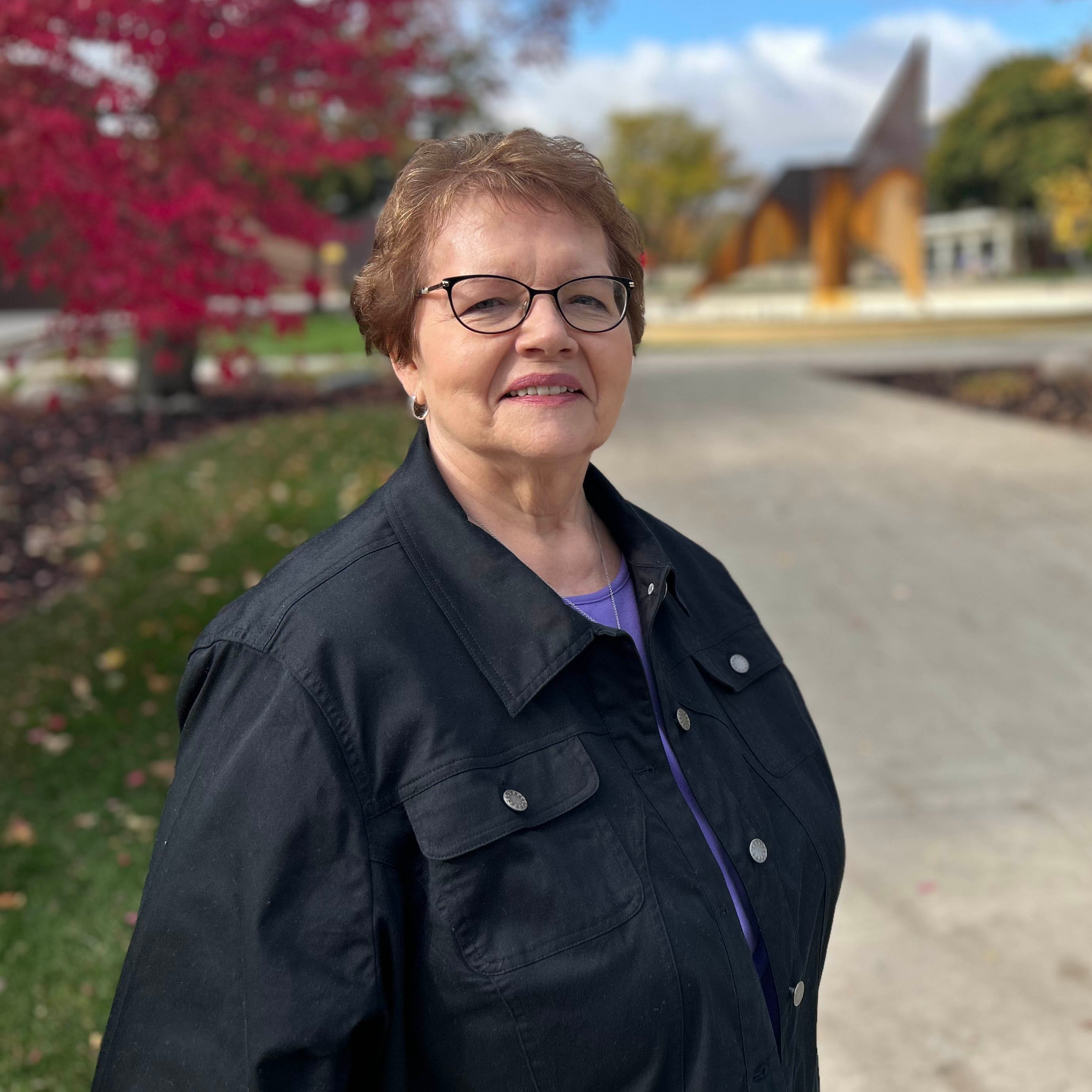 Jan Gostonczik poses outside with the campus and fall colors in the background. She is smiling at the camera.