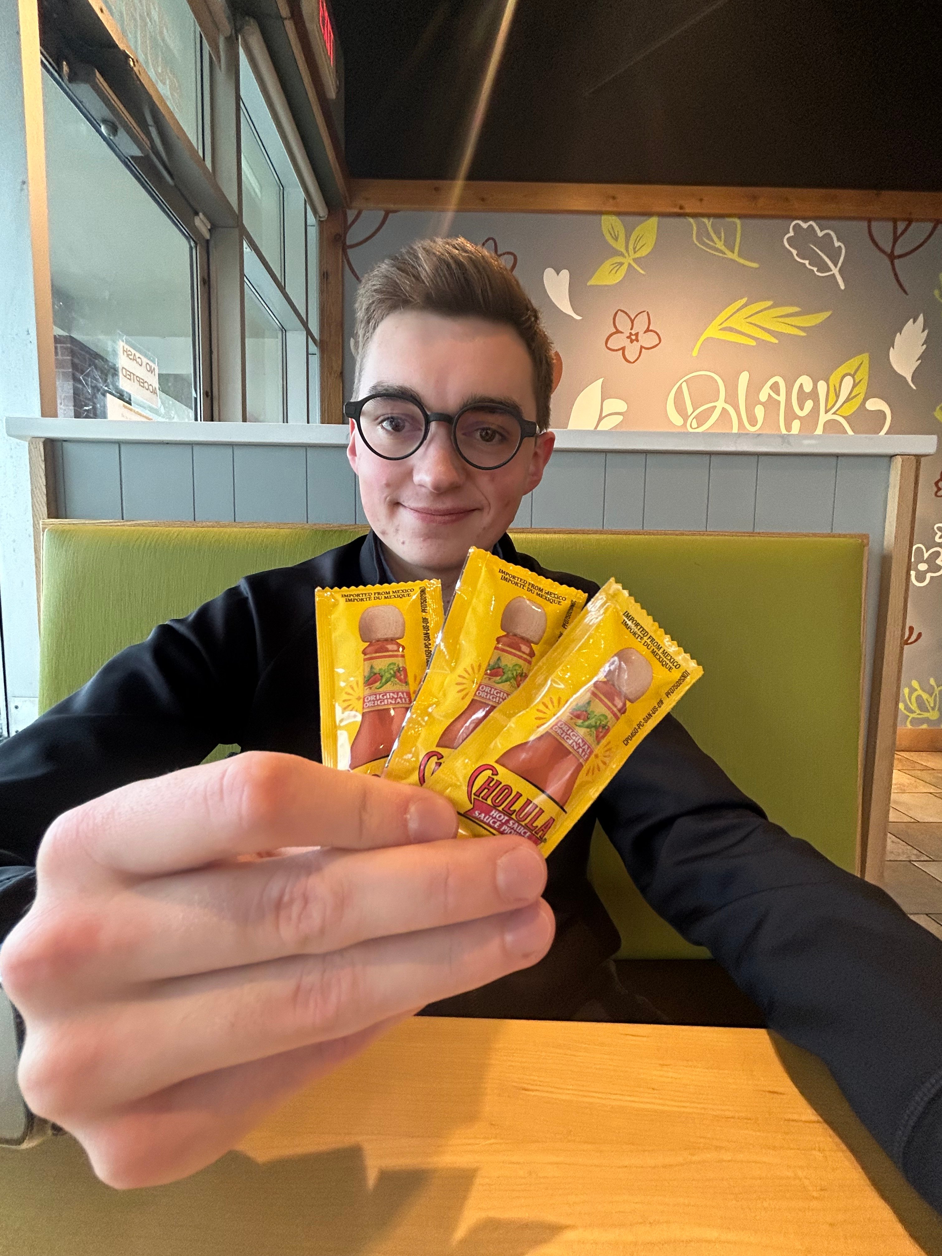 Brandon sitting in a booth at a restaurant holding up packets of hot sauce and smiling at the camera. 
