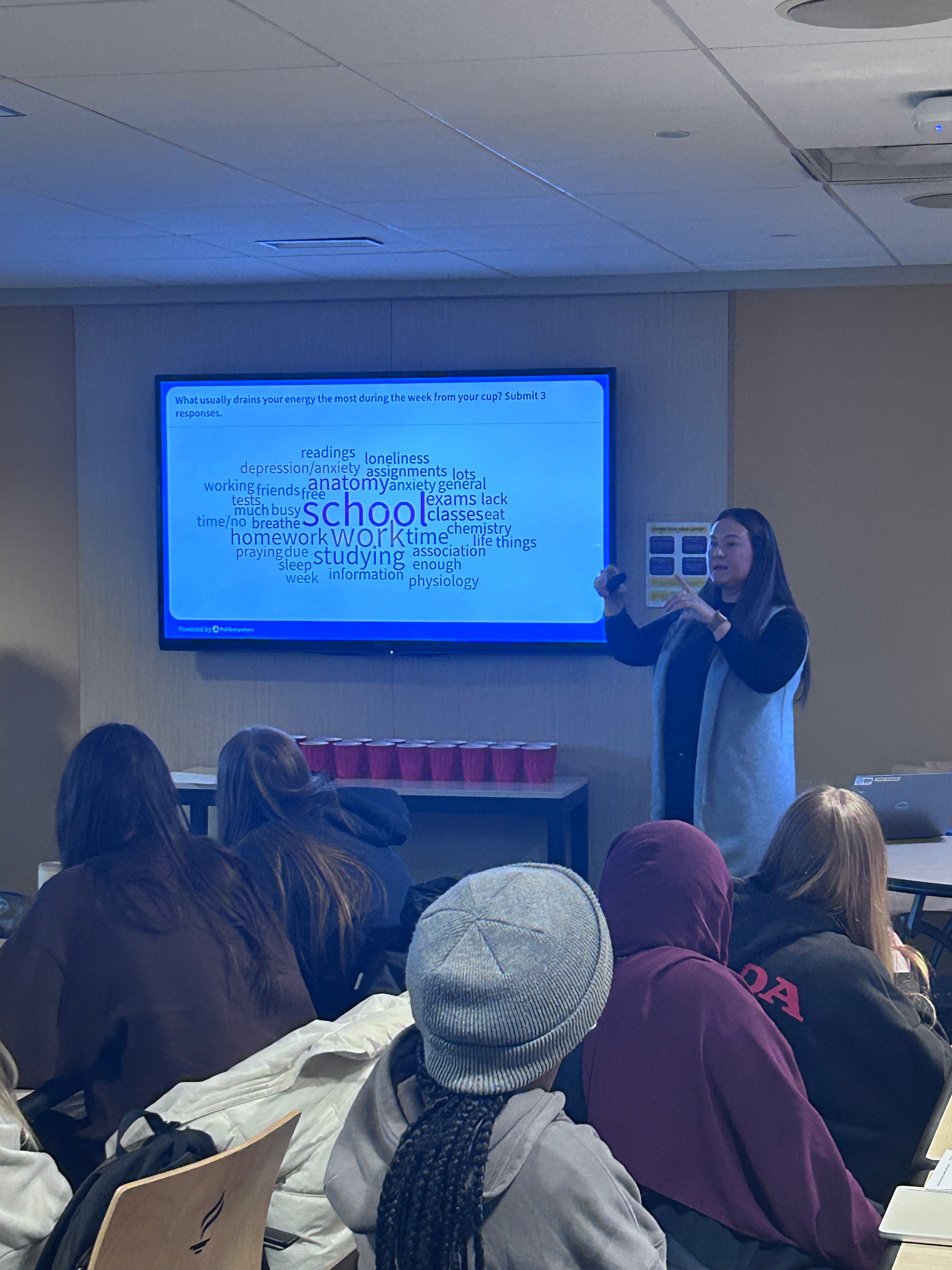Justine Schultz, health educator, presenting to a room full of students. The PPT slide on the wall lists out what usually drains your energy the most during the week. Such as School, work, studying, life things