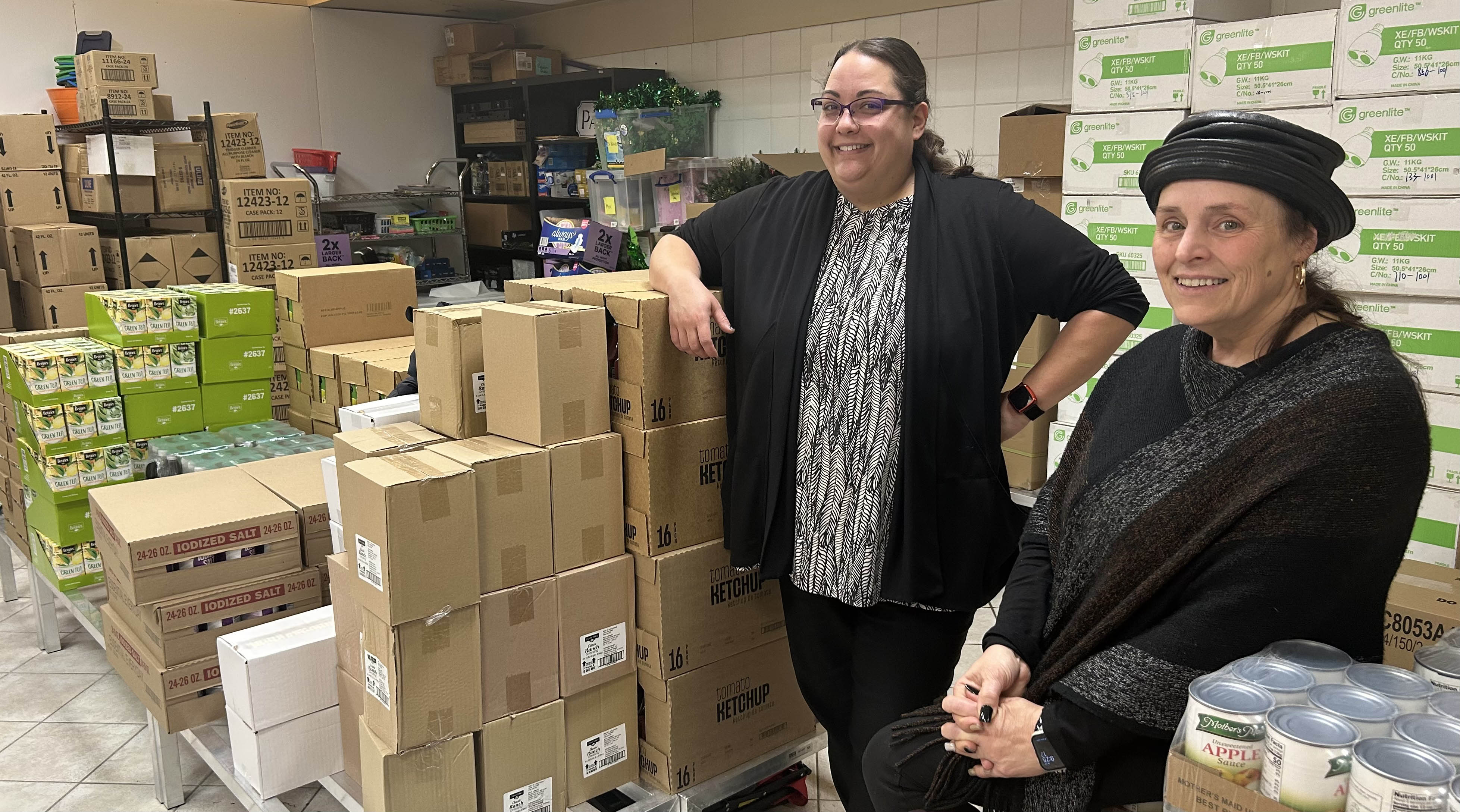 Two female MSU staff advisors to the Food Pantry posing with stacks of food boxes in the background.