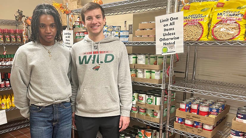 Two students standing in front of the food shelves where students can shop for food. There are bottles of sauce, mustard, soup and more on the shelves. Both students are smiling.