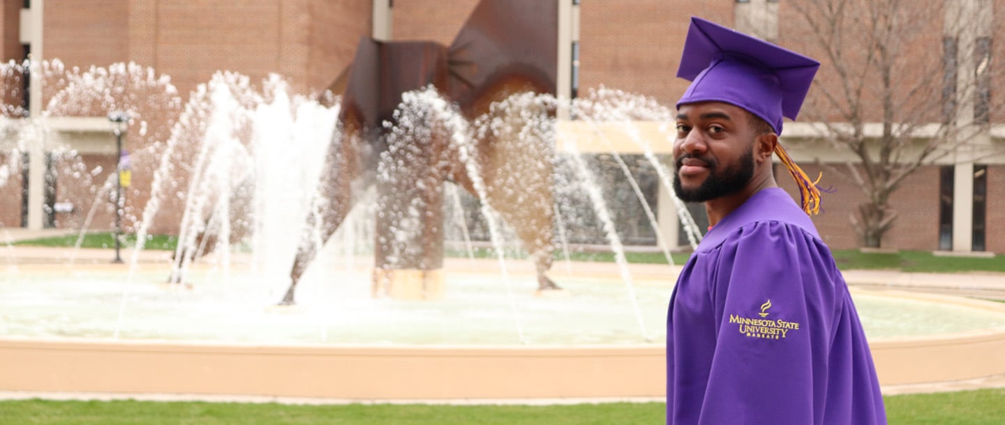 A student in a purple graduation gown and hat, standing outside by the fountain between the Centennial Student Union building and Memorial Library