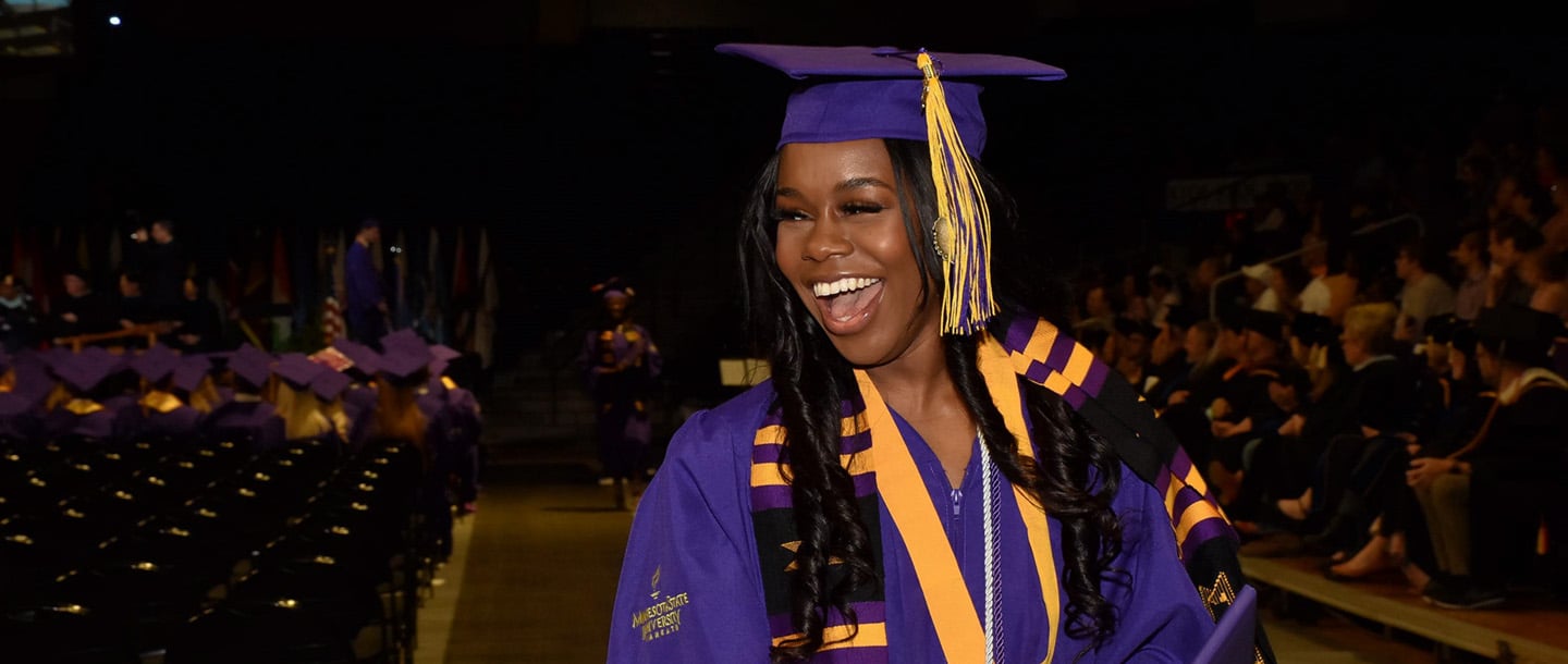 A student smiling and walking in a graduation ceremony wearing a purple graduation gown and hat