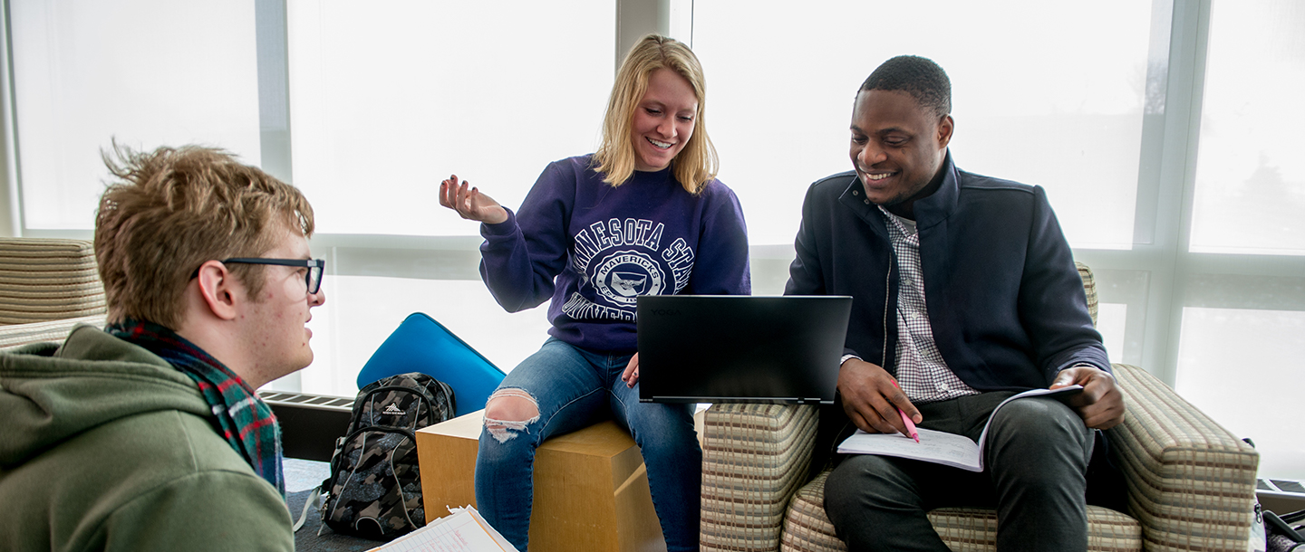 Three students sitting in the lounge studying together with a laptop and notebooks
