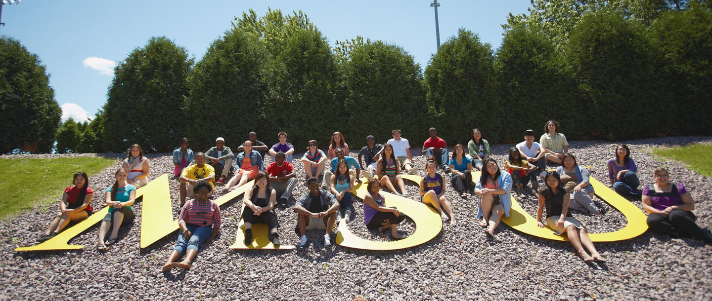 A group of students posing and sitting on the yellow 'MSU' letters at Minnesota State University next to the football stadium