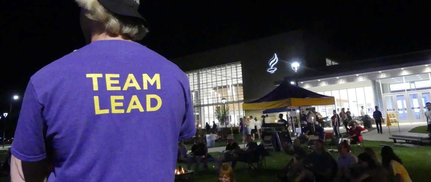 An individual at an event right by the university dining wearing a purple colored shirt which says team lead from the back