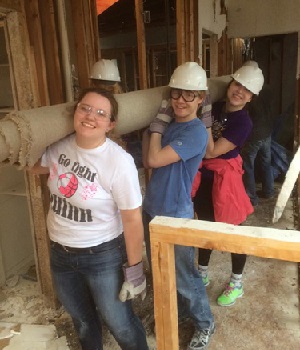 Three student volunteers posing at a building construction site while holding rolled up carpet