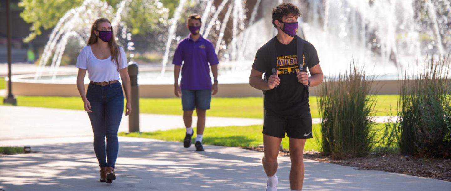 Three students wearing masks walking in front of the fountain towards Memorial Library