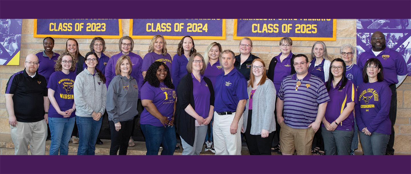 A group of university advising professionals in purple shirts standing in front of a stone brick fireplace.