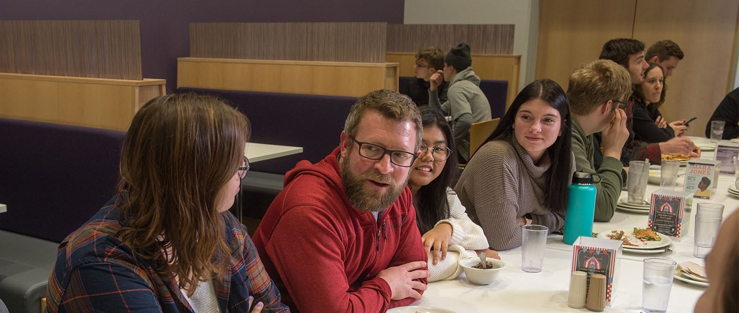 A group of students, at a table in the dining center, chatting with a faculty member
