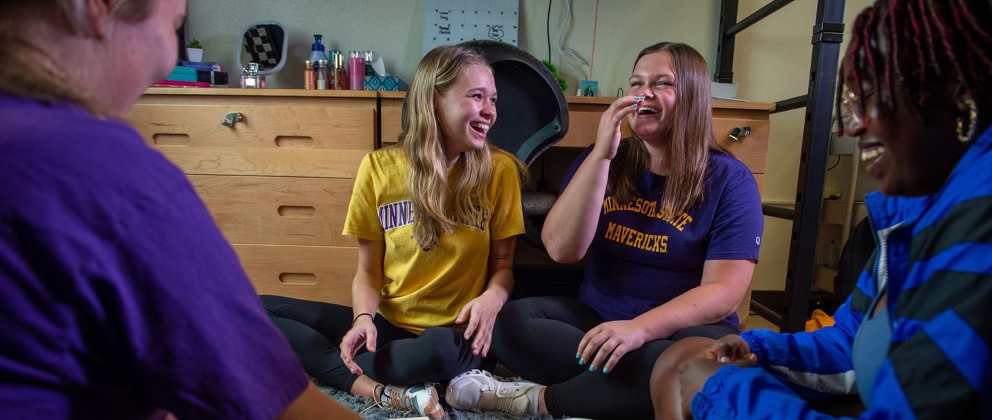 Four students living on campus sitting on the floor of their dorm room and laughing with their roommates