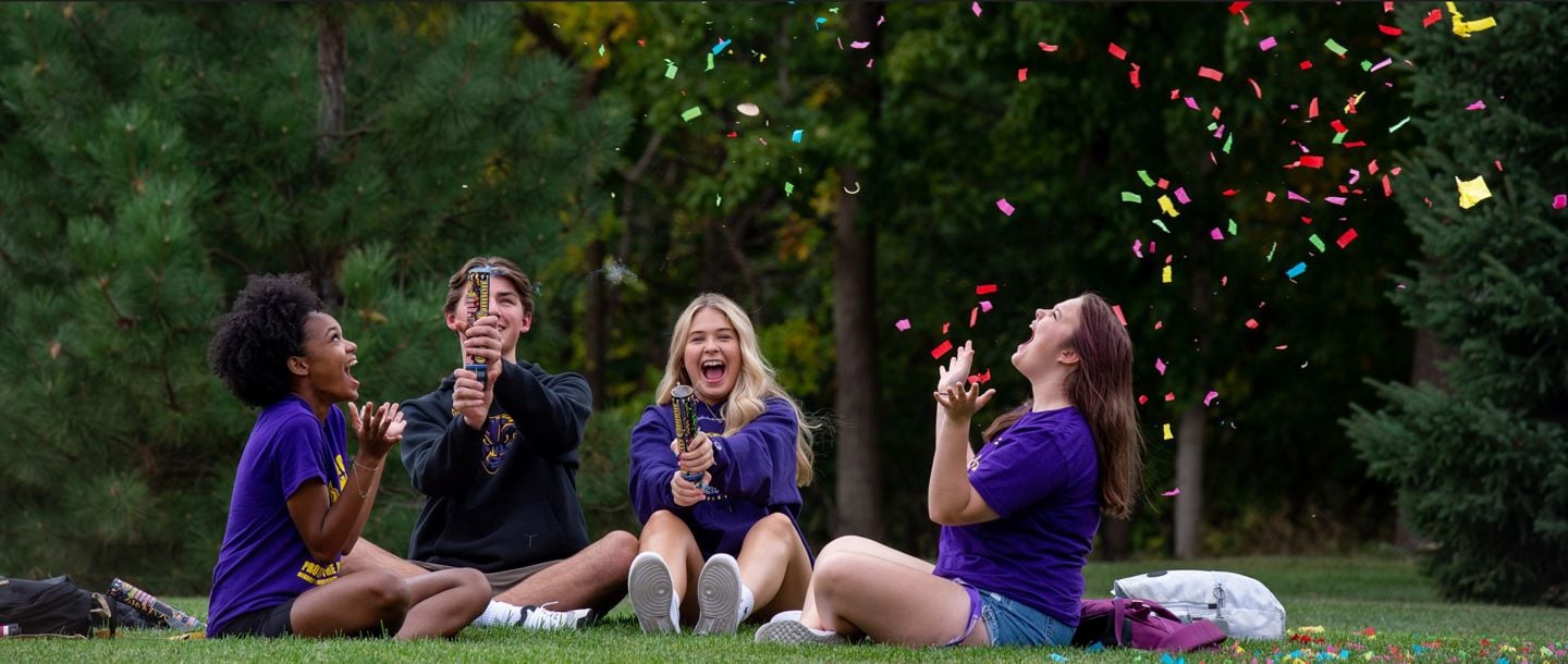 Four students hanging out together outside sitting on the campus lawn, two of them are using confetti canons to shoot confetti up in the air