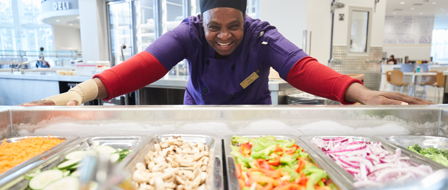 A University Dinning Center cook smiling behind the counter with various food options