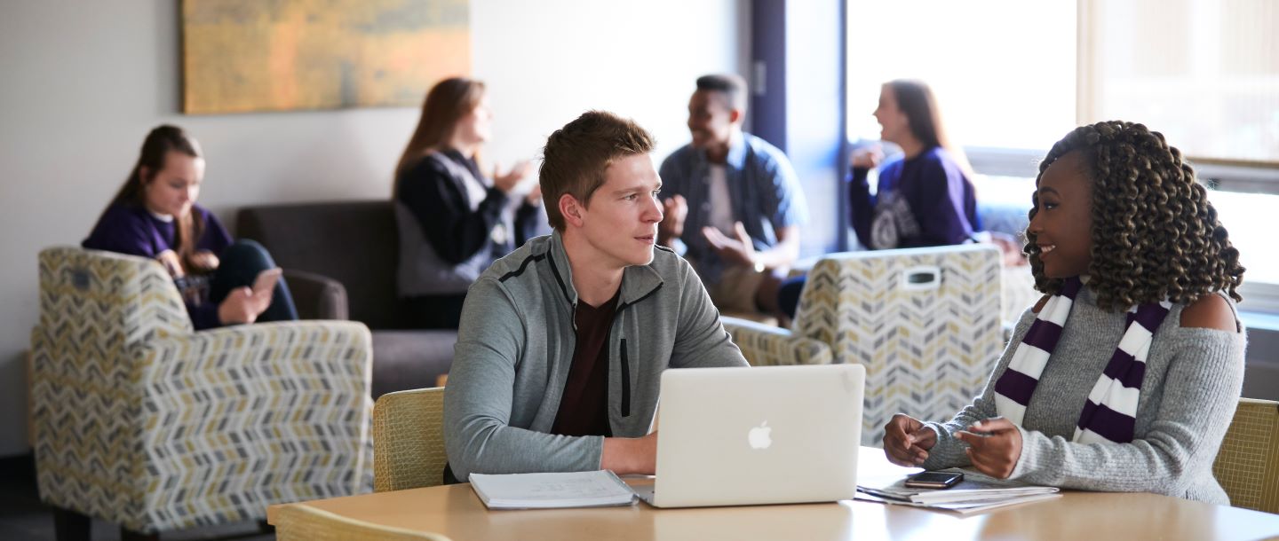 Two students in a discussion at table with laptop and a notebook