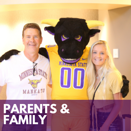 Parents and family photo with a student and her father standing in the hallway on campus posing for a photo with Stomper, the Maverick mascot