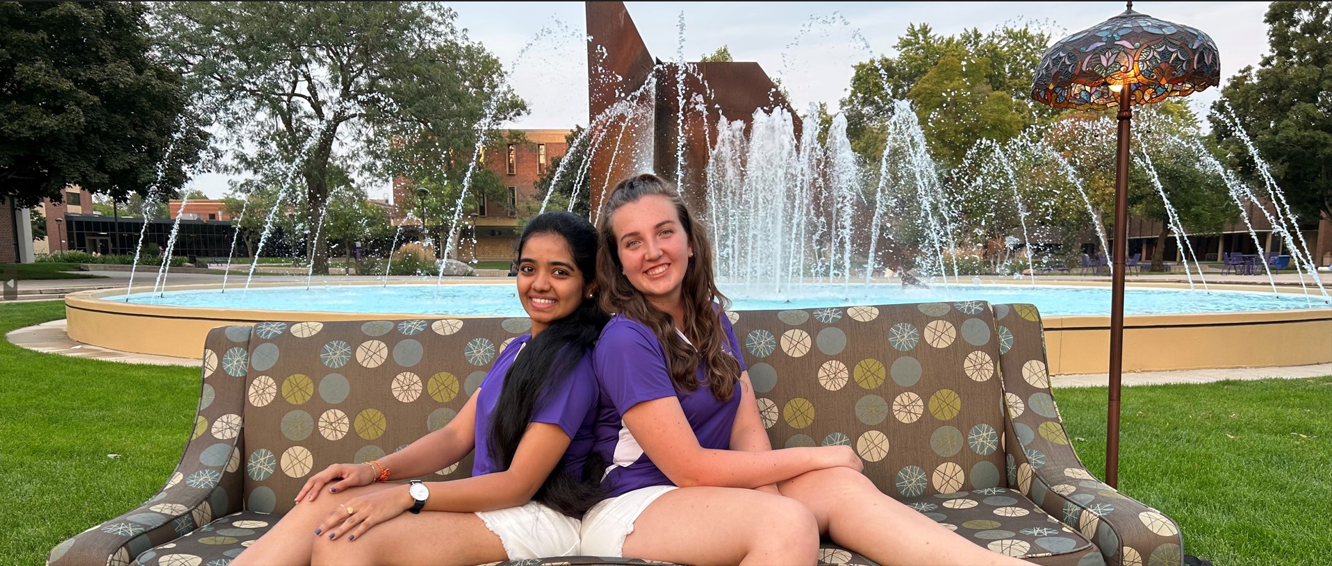 two women sitting on a bench with a fountain in the background