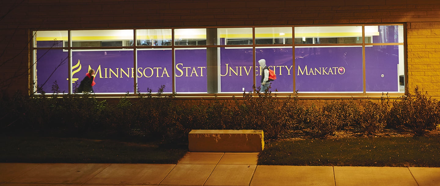 Two students walking indoors opposite directions in the hallwayby with the Minnesota State University Mankato logo in the background