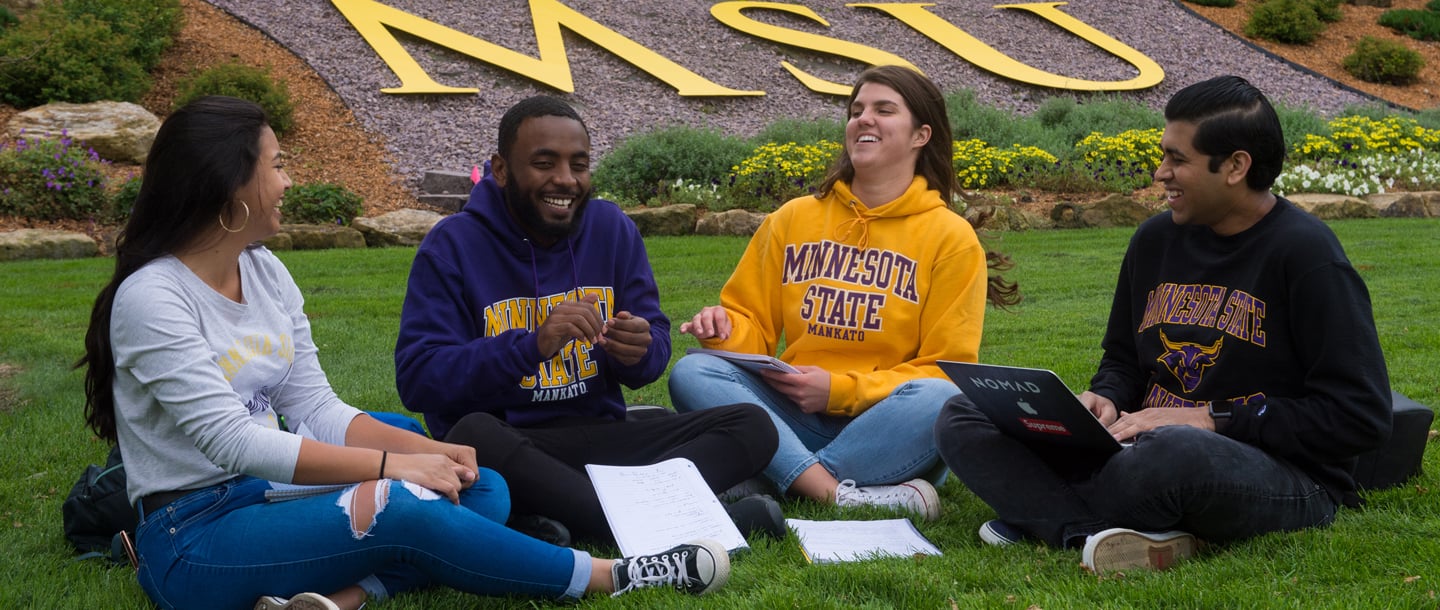 Four students sitting outside on the lawn with the yellow letters "MSU" and flowers in the background