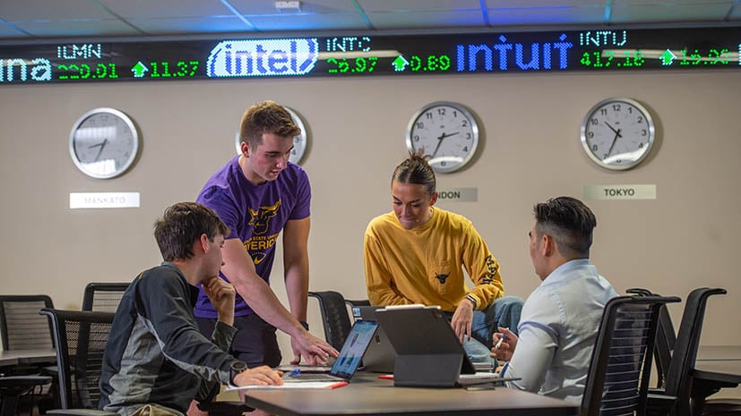 four students sitting in the finance room with the stock market ticker in the background