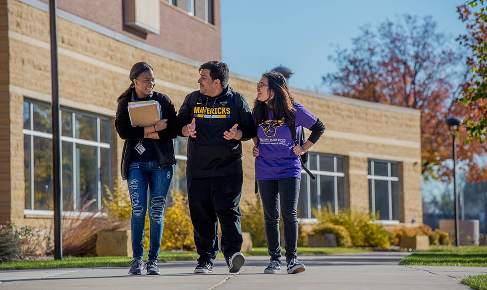 Three students walking on campus in their Maverick gear, backpacks, and talking and laughing